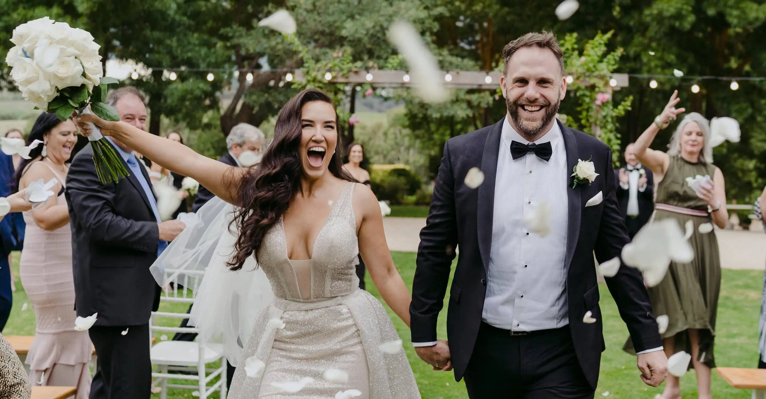 Newlyweds holding hands beneath falling white petals as guests celebrate