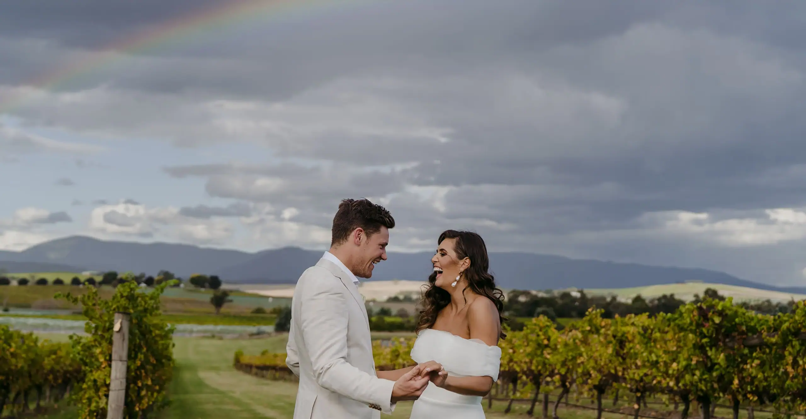 Couple laughing and holding hands beside vineyard rows beneath a cloudy sky