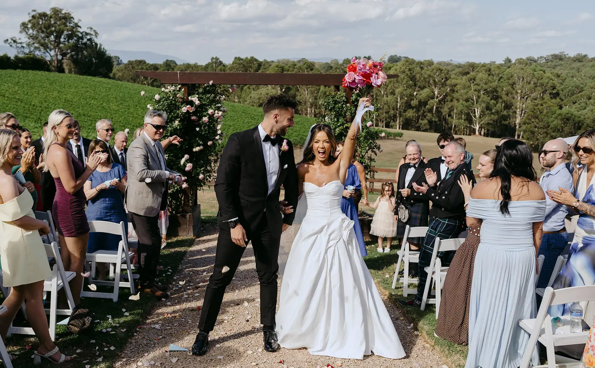 Bride raising a pink bouquet while walking with the groom between applauding ceremony guests.